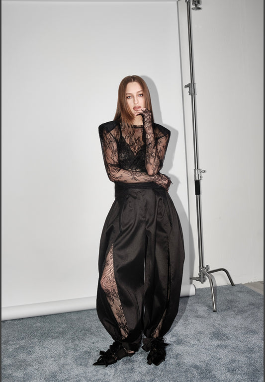 Outfit Ginevra elegante e sensuale composto da maglia in pizzo trasparente e pantalone nero a vita alta. Il pantalone presenta una linea fluida con spacco laterale e dettagli con fiocchi alla caviglia,